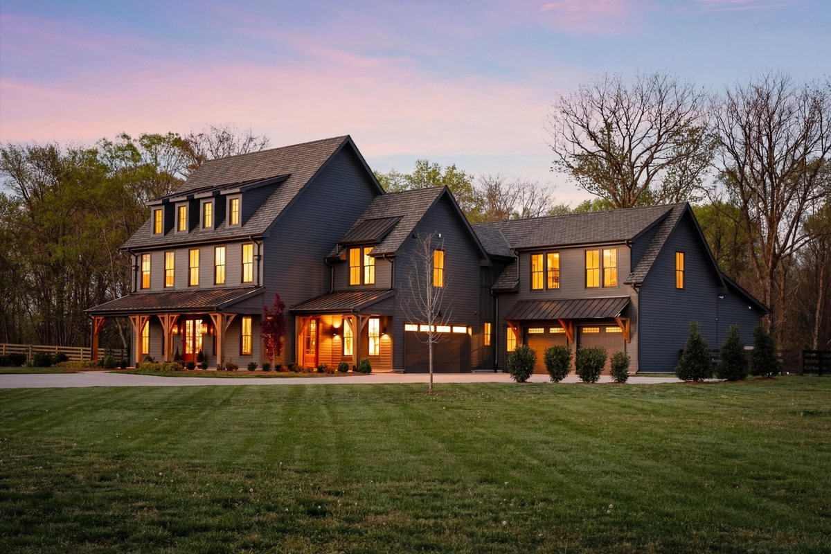 Dark modern farmhouse with warmly lit windows, timber porch posts and a wide lawn at dusk